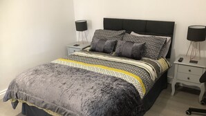 A neatly made bed with a dark headboard, a patterned bedspread, and multiple pillows. There are two bedside tables with lamps and a small drawer. - Catherines Court (Solihull)
