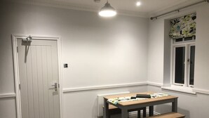 A room with a white door, a window with a floral blind, a table with a bench, and a ceiling light. - Catherines Court (Solihull)