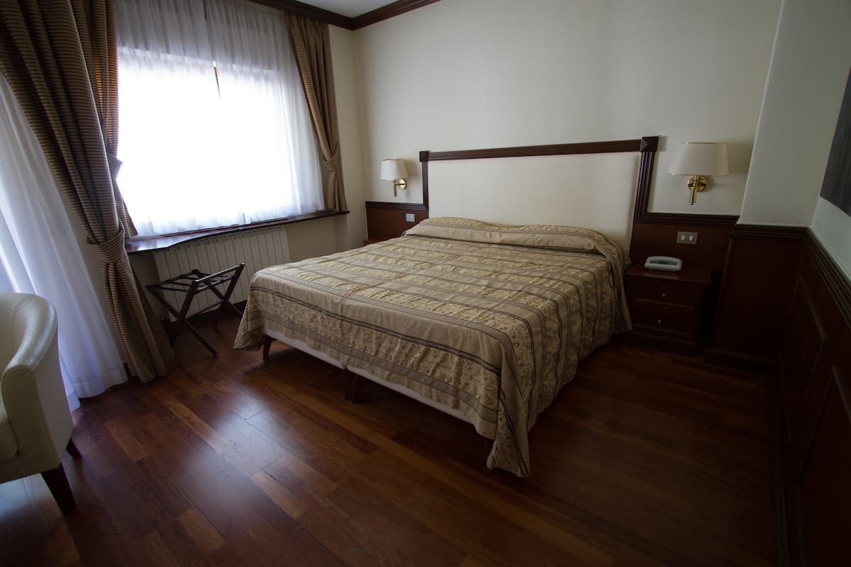 Una cama bien hecha con una colcha estampada, una mesita de noche de madera con una lámpara, una silla y una ventana con cortinas.