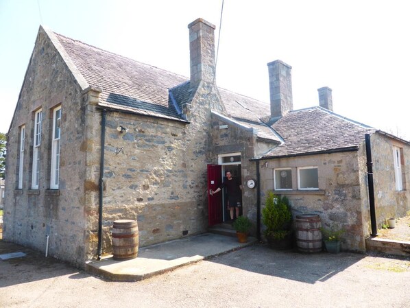 A stone building with a red door, two chimneys, and a small porch area with barrels and potted plants.