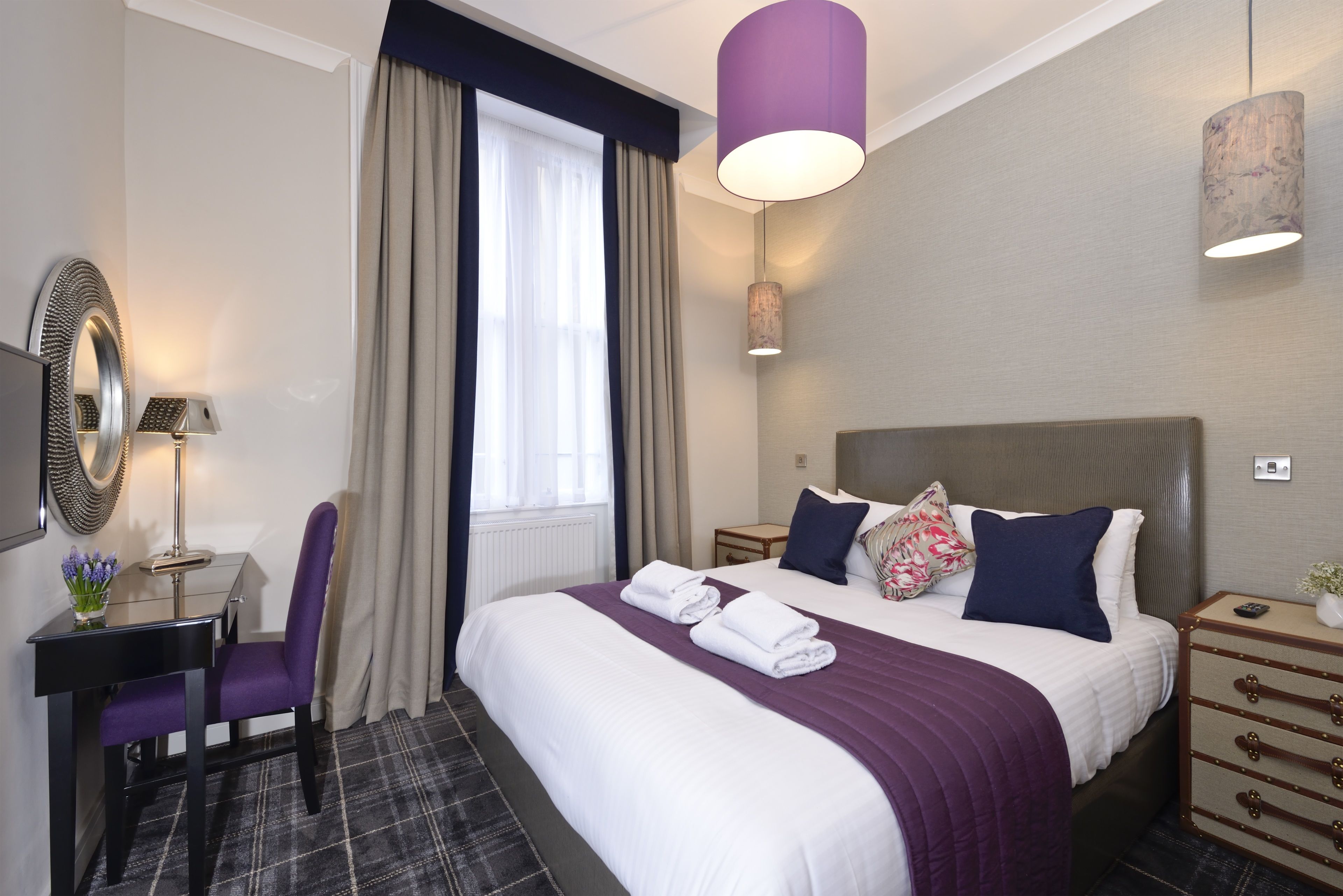 A hotel room with a large bed, a desk with a chair, a mirror, and a window with curtains.