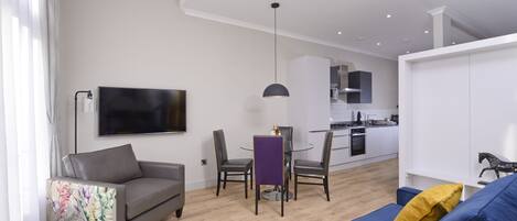 A modern living room with a sofa, a dining table with chairs, a TV mounted on the wall, and a kitchen in the background.