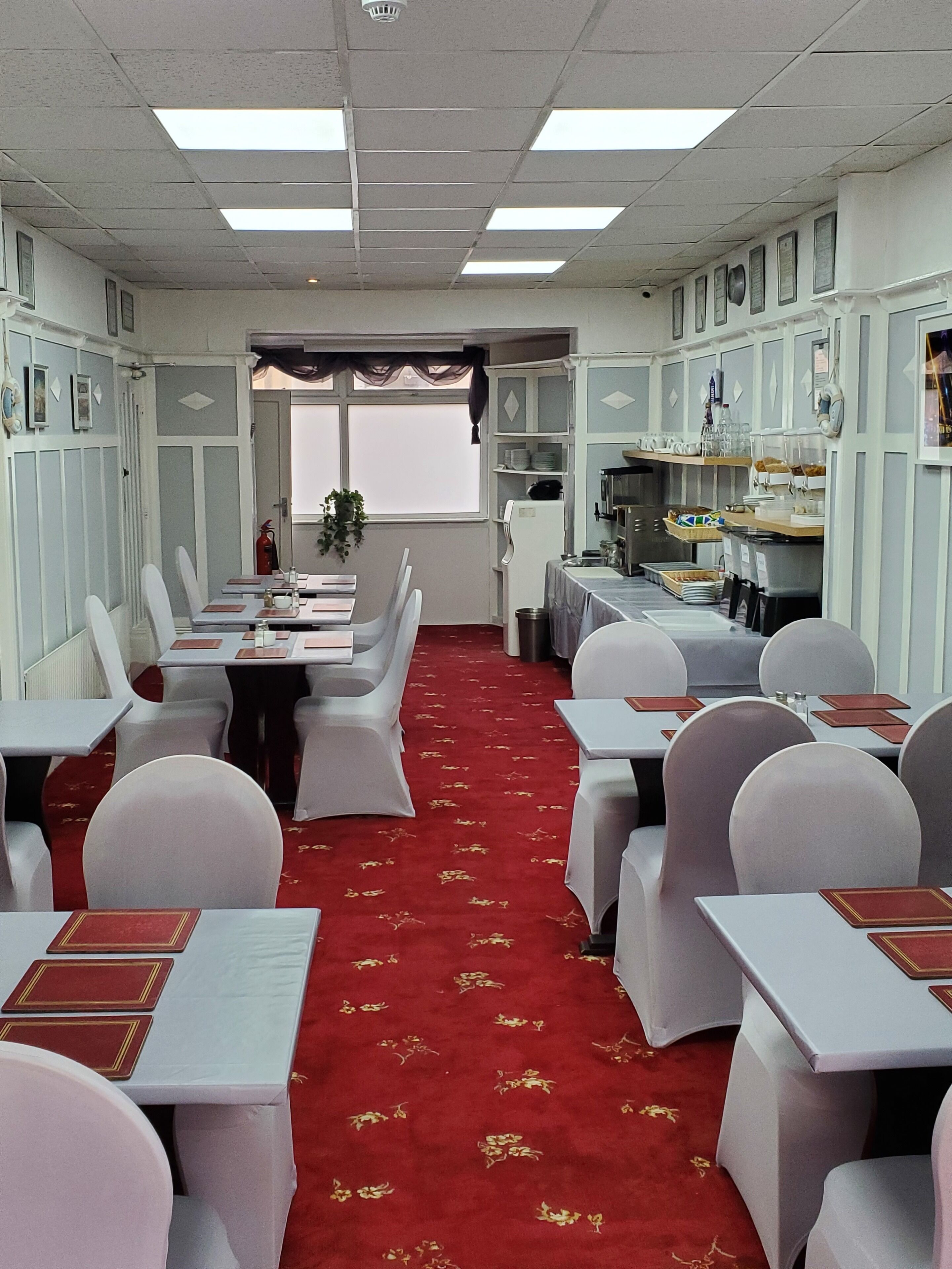 Un salón de conferencias con mesas y sillas blancas, una alfombra roja y una zona de cocina con estanterías y electrodomésticos.