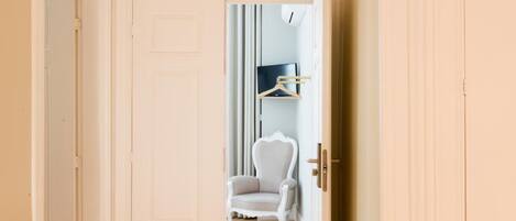 A hallway with wooden flooring and white doors leading to a room with a chair and a coat rack.