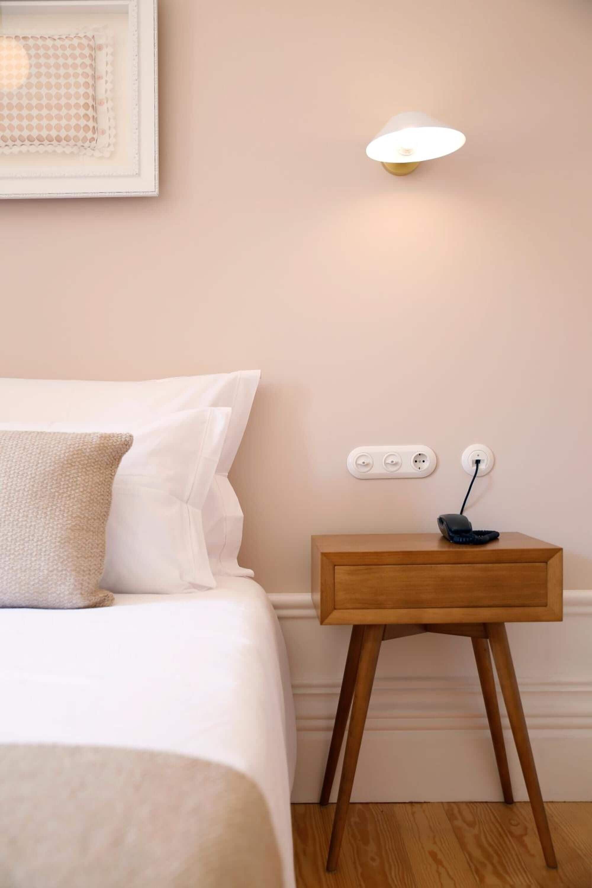 A neatly made bed with white linens and a single pillow, a bedside table with a lamp, and a power outlet on the wall.