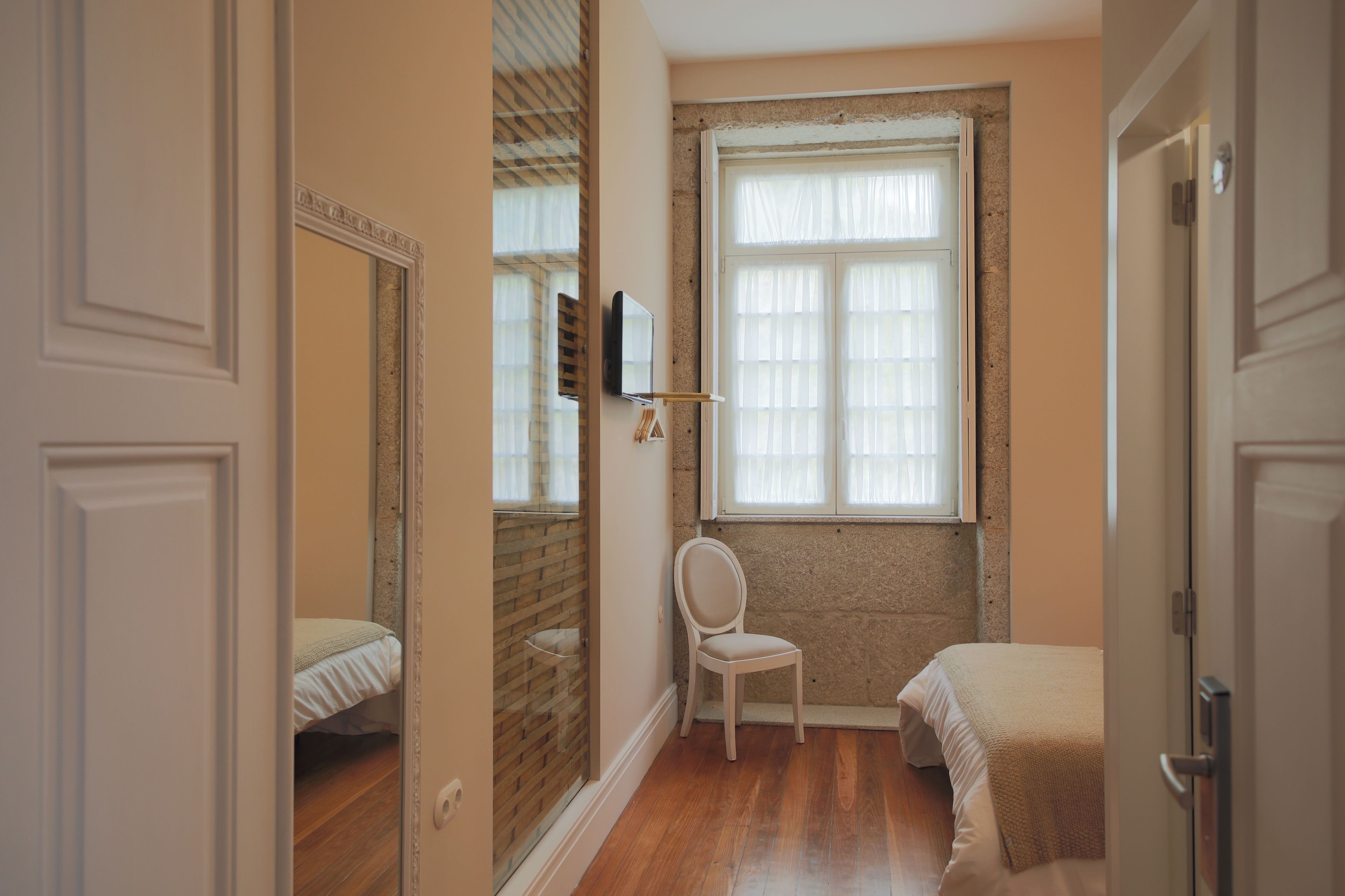 A narrow room with a wooden floor, a white chair, a bed with a beige blanket, a window with sheer curtains, and a mirror on the wall.