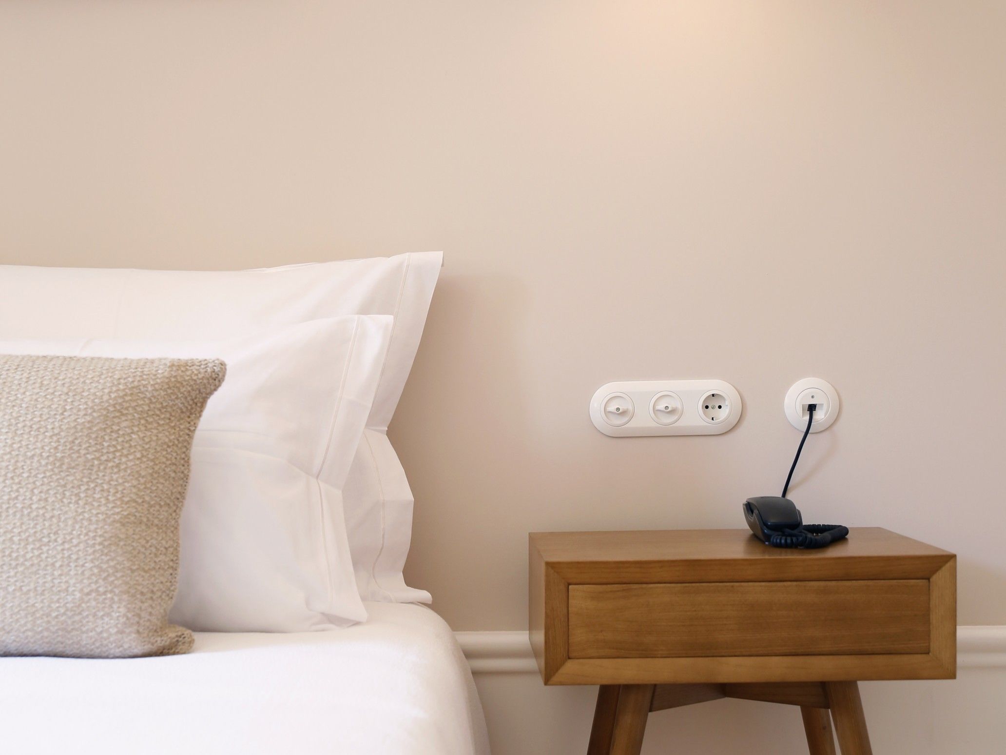 A neatly made bed with white linens and a textured pillow, a bedside table with a phone, and a wall socket.