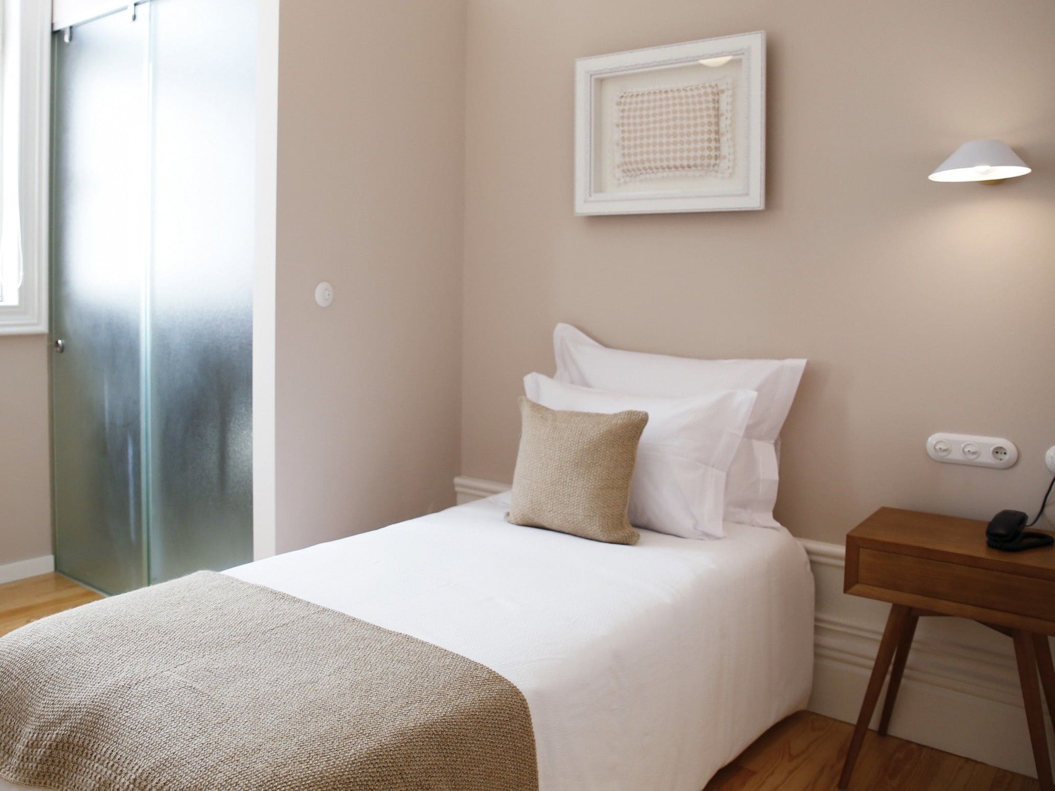 Una cama bien hecha con sábanas blancas y una manta color beige, una mesita de noche con una lámpara y una obra de arte enmarcada en la pared.
