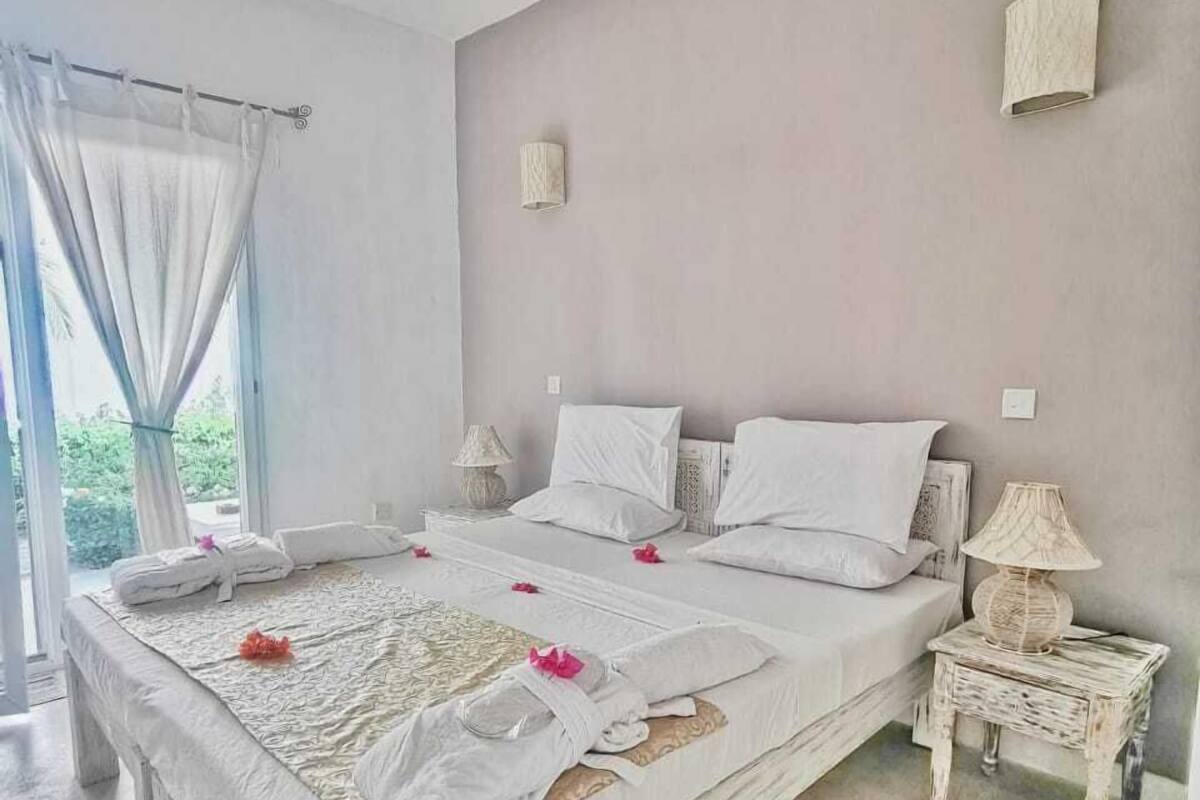 A bedroom with a large bed, two bedside tables, a window with curtains, and a wall-mounted lamp.