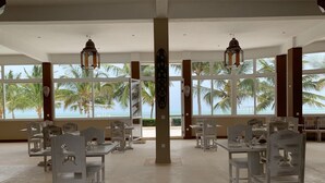 A spacious dining area with white tables and chairs, large windows offering a view of palm trees and the ocean. - Life Resort St.Thomas Royal Palm (Watamu)