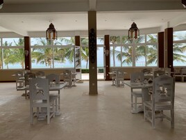 A spacious dining area with white tables and chairs, large windows offering a view of palm trees and the ocean.