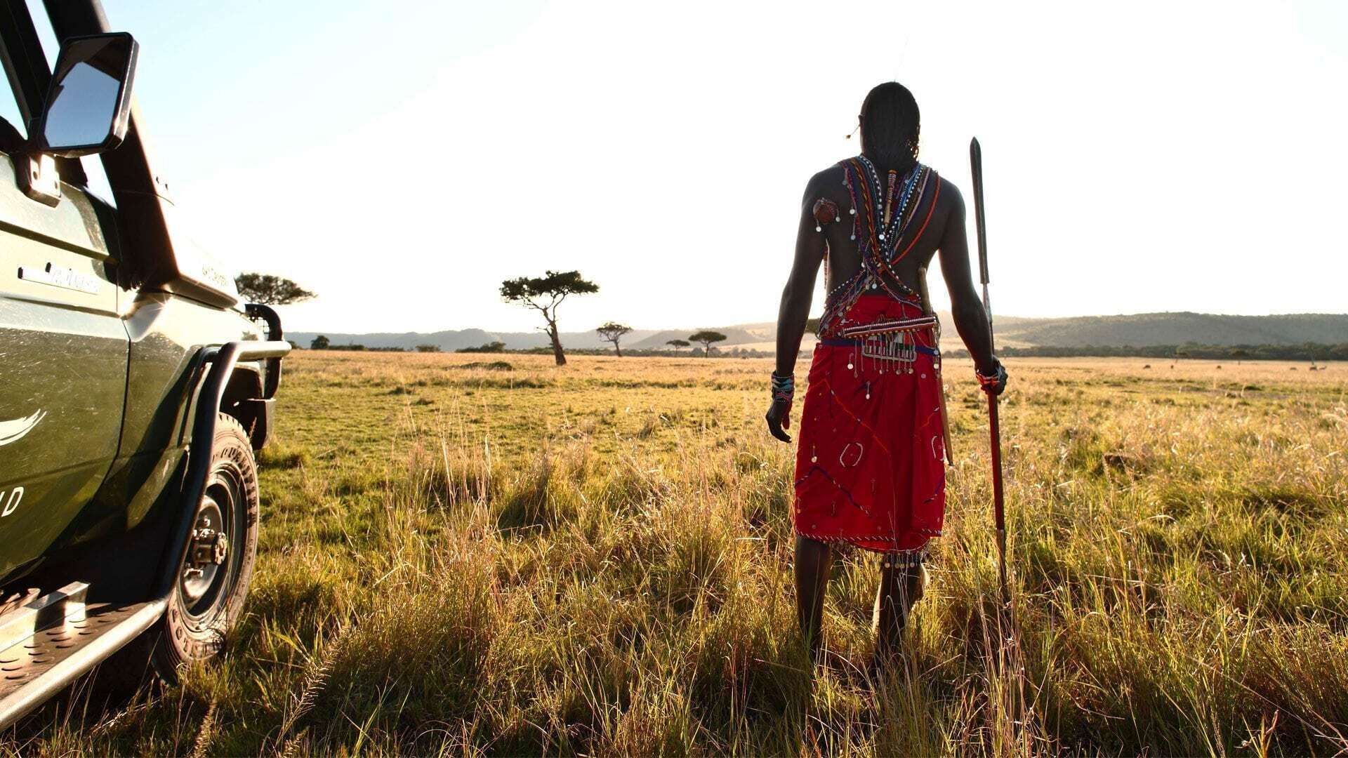 A safari vehicle parked in a grassy field with a person in traditional attire standing nearby.