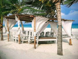 A beach cabana with white chairs and a thatched roof, overlooking the ocean.