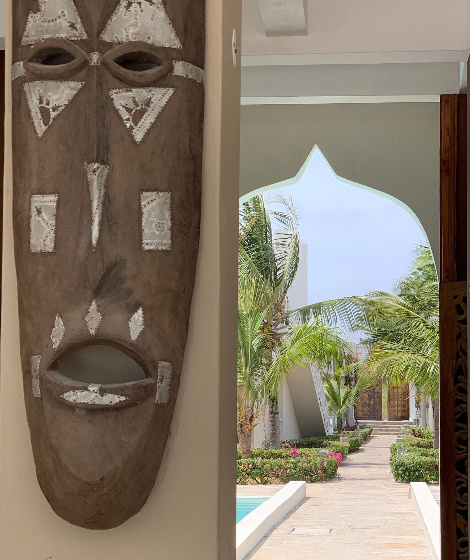 Een houten masker met geometrische patronen en een pad dat leidt naar een gebouw met een bijzondere boog en palmbomen.