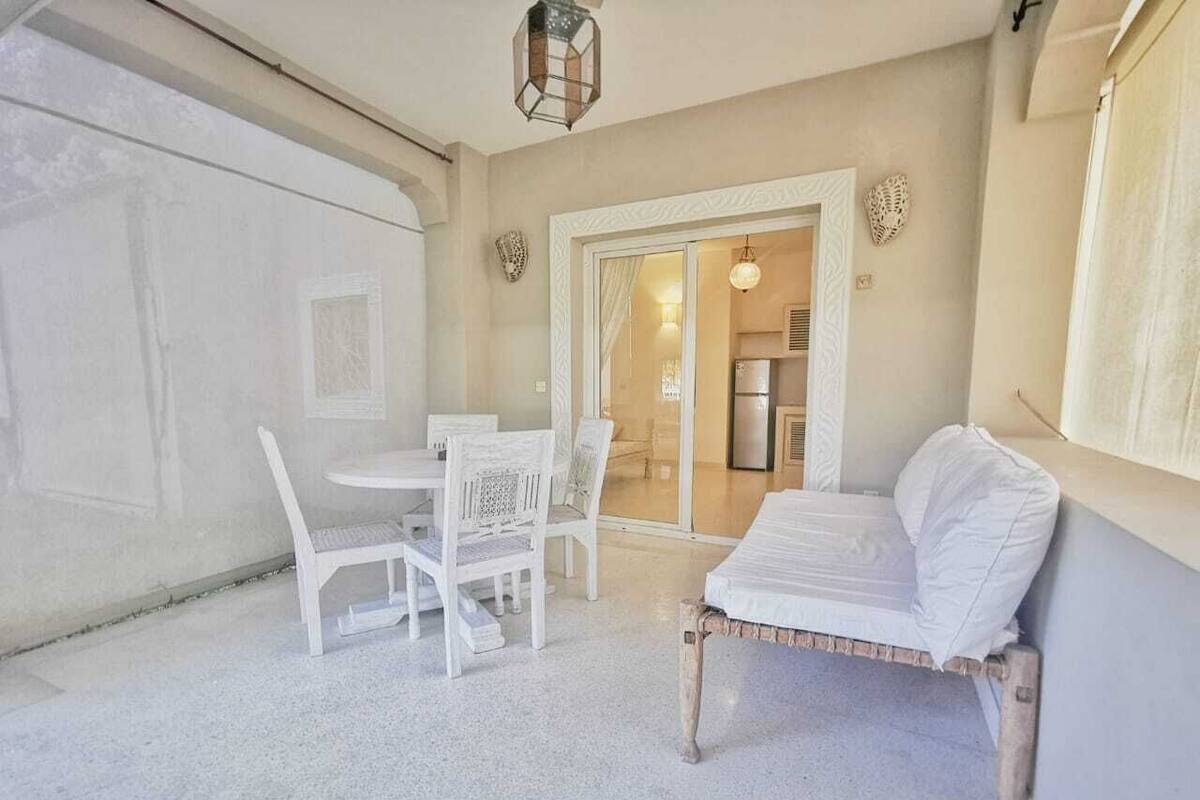 A room with a white table and chairs, a wooden bench, and a door leading to a kitchen area.