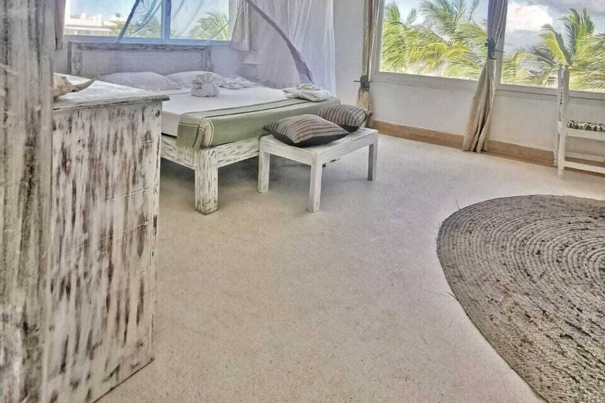A bedroom with a bed, a small table, a bench, a large window, and a view of palm trees.