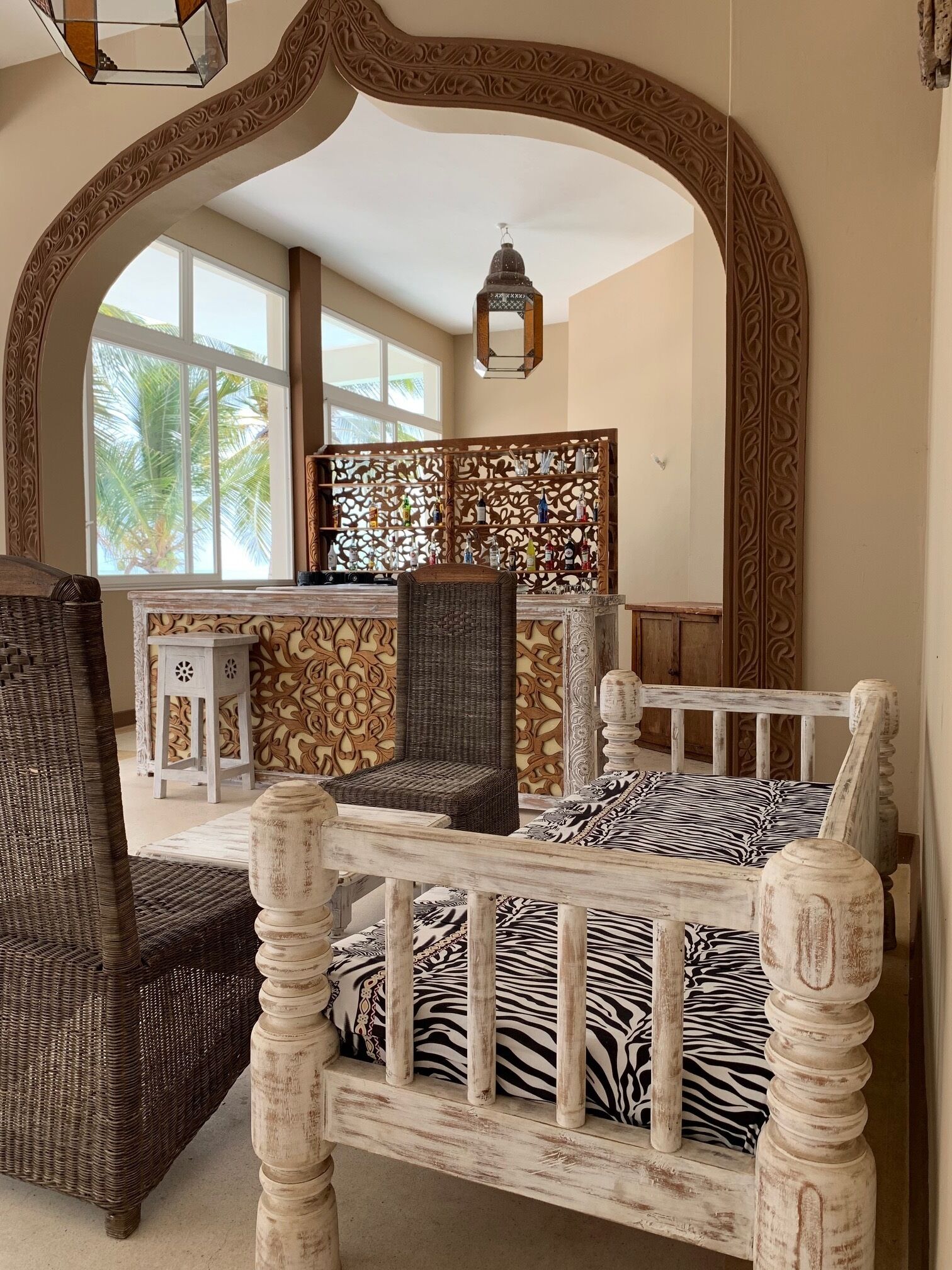 A room with a bed featuring a zebra print, a wicker chair, a decorative wall panel, and a view of palm trees through large windows.