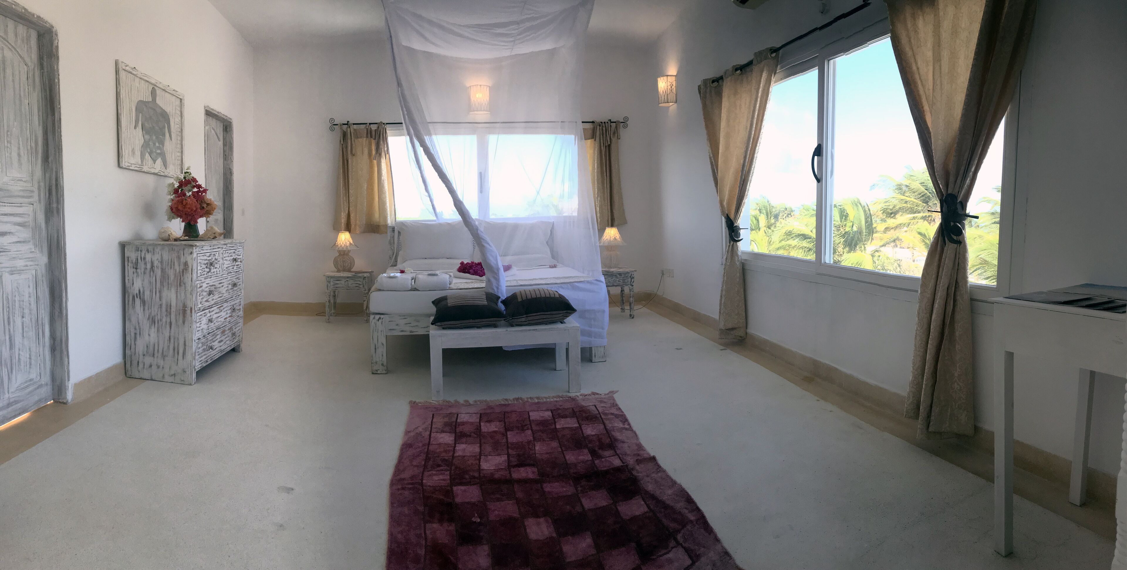Un dormitorio con cama con dosel, una mesita de noche con lámpara, un jarrón decorativo y vistas a palmeras a través de la ventana.