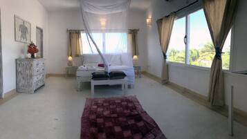 Un dormitorio con cama con dosel, una mesita de noche con lámpara, un jarrón decorativo y vistas a palmeras a través de la ventana.