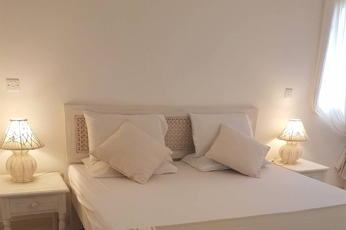 A neatly made bed with white linens and pillows, two bedside tables with lamps, and a plain wall with a window.