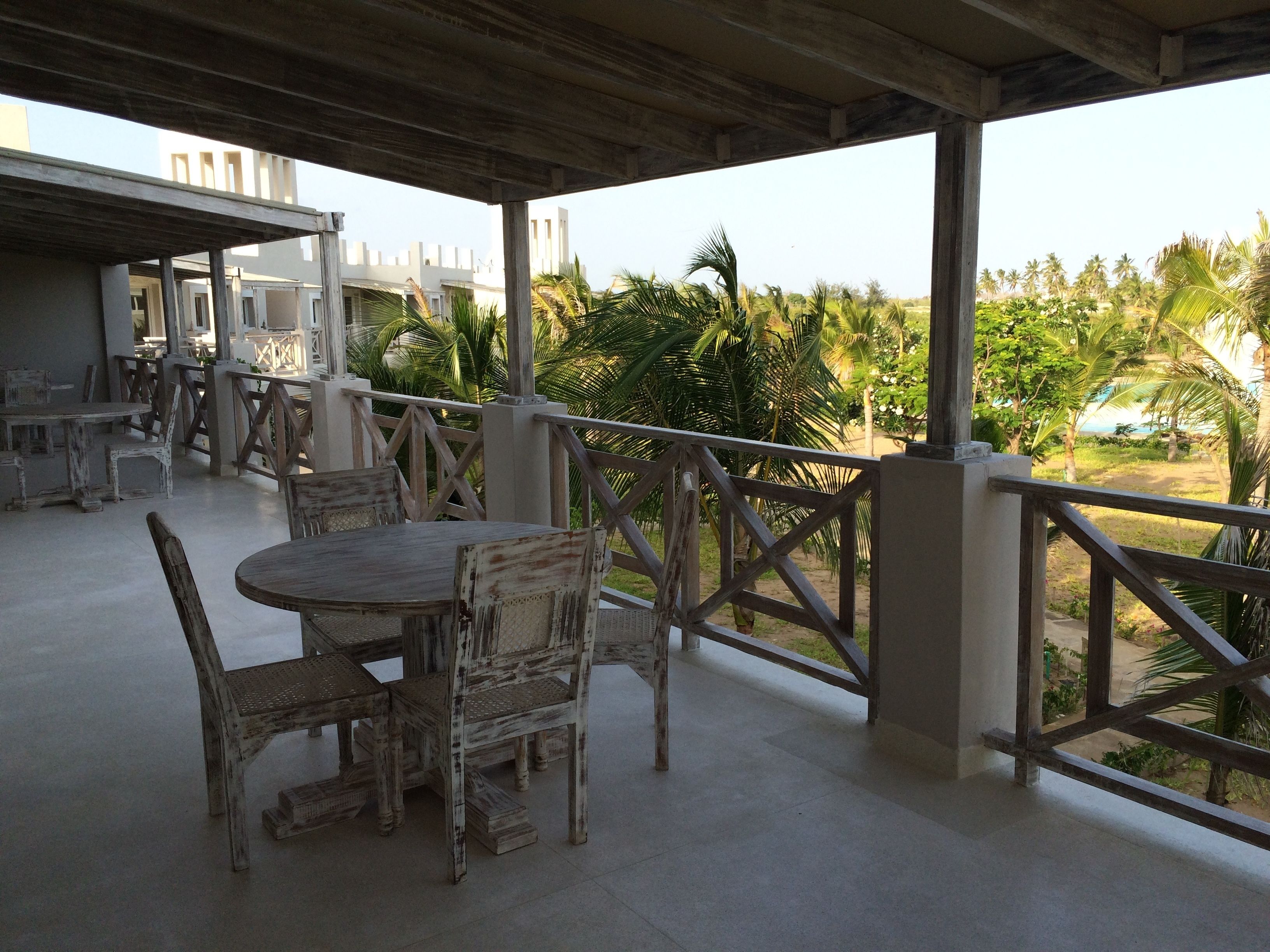 A covered outdoor seating area with a table and chairs, a railing, and a view of palm trees and buildings in the background.