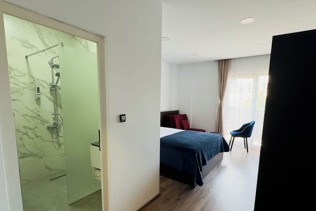 A modern hotel room with a glass shower door, a bed with a blue bedspread, a wooden floor, and a window with curtains.