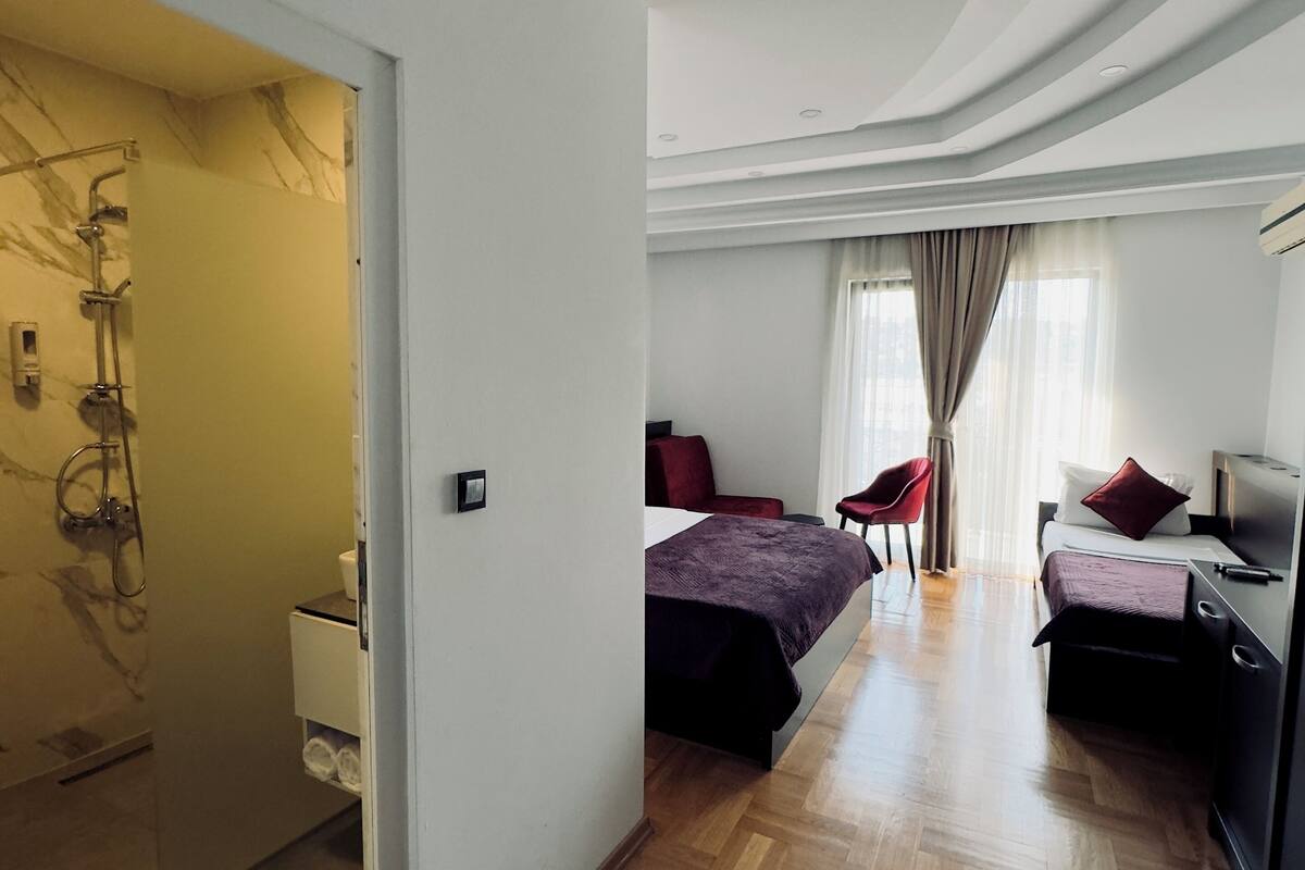 A hotel room with a wooden floor, a bed with a purple bedspread, a desk with a chair, a shower area, and a bathroom with a marble wall.
