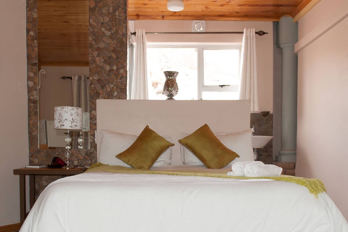 A bedroom with a bed, two pillows, a nightstand with a lamp, a window with curtains, and a stone wall.