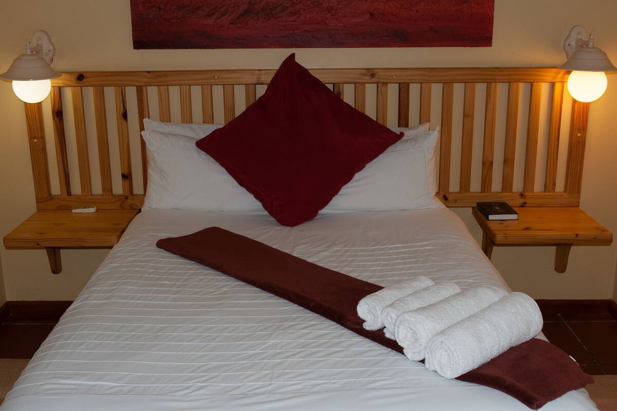 A bed with white linens, a red throw, and white towels. There are wooden headboards and nightstands with lamps.
