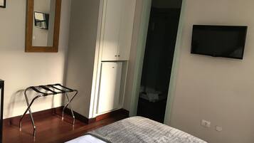 Un dormitorio con cama, escritorio de madera, espejo, armario, televisor y baño visible a través de una puerta.