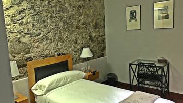 Un dormitorio con pared de piedra, cabecera de madera, cama con ropa blanca, mesita de noche con lámpara, pequeña mesa con silla y dos cuadros enmarcados en la pared.