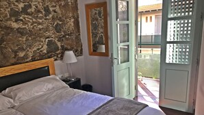 Un dormitori amb paret de pedra, un llit amb roba de llit blanca, una nitzeta amb una làmpada i vista a un pati a través d'una porta oberta.