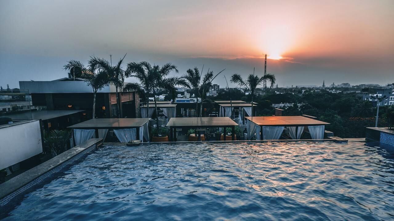 A rooftop pool area with tables and chairs, palm trees, and a sunset view.