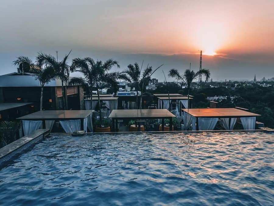 A rooftop pool area with tables and chairs, palm trees, and a sunset view.