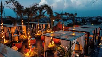 An outdoor seating area with tables and chairs, surrounded by potted plants and draped fabric.