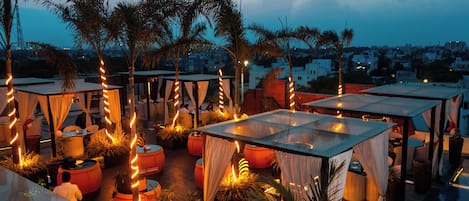 An outdoor seating area with tables and chairs, surrounded by potted plants and draped fabric.