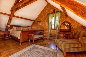 Ein Schlafzimmer mit schräg verlaufender Decke, Holzbalken, einem Bett, einem Sessel, einer Kommode und einem Spiegel.