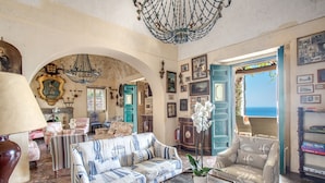 Un vaste salon agrémenté d’un lustre, d’un canapé, de fauteuils et offrant une vue sur la mer grâce à une porte ouverte.