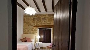 Un couloir étroit mène à une chambre dotée d’un mur en pierre, de poutres en bois, d’un lit, d’une table de chevet et d’une lampe.