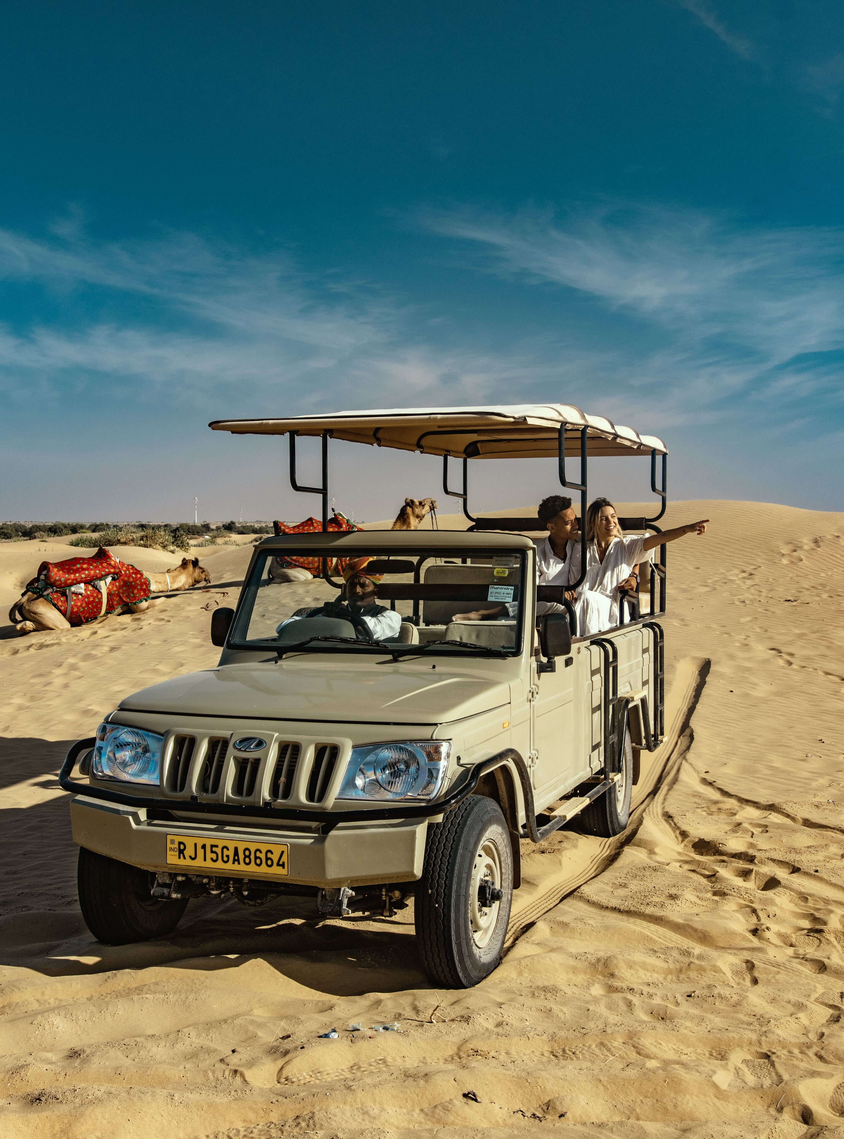 A safari vehicle parked on a sandy dune with camels in the background.