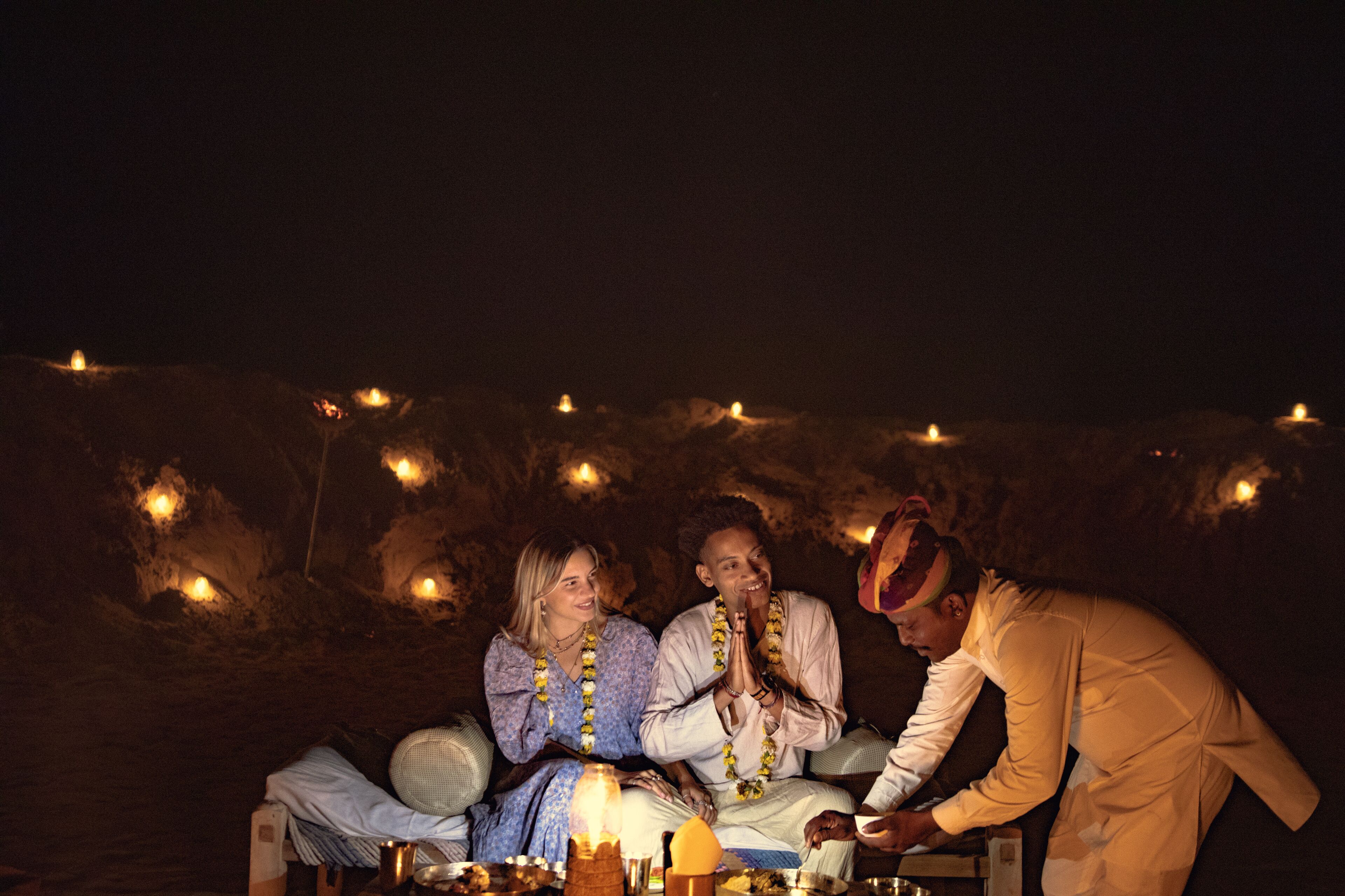 Un grupo de personas sentadas alrededor de una mesa con una vela en el centro, en un paisaje rocoso y oscuro con pequeñas luces al fondo.