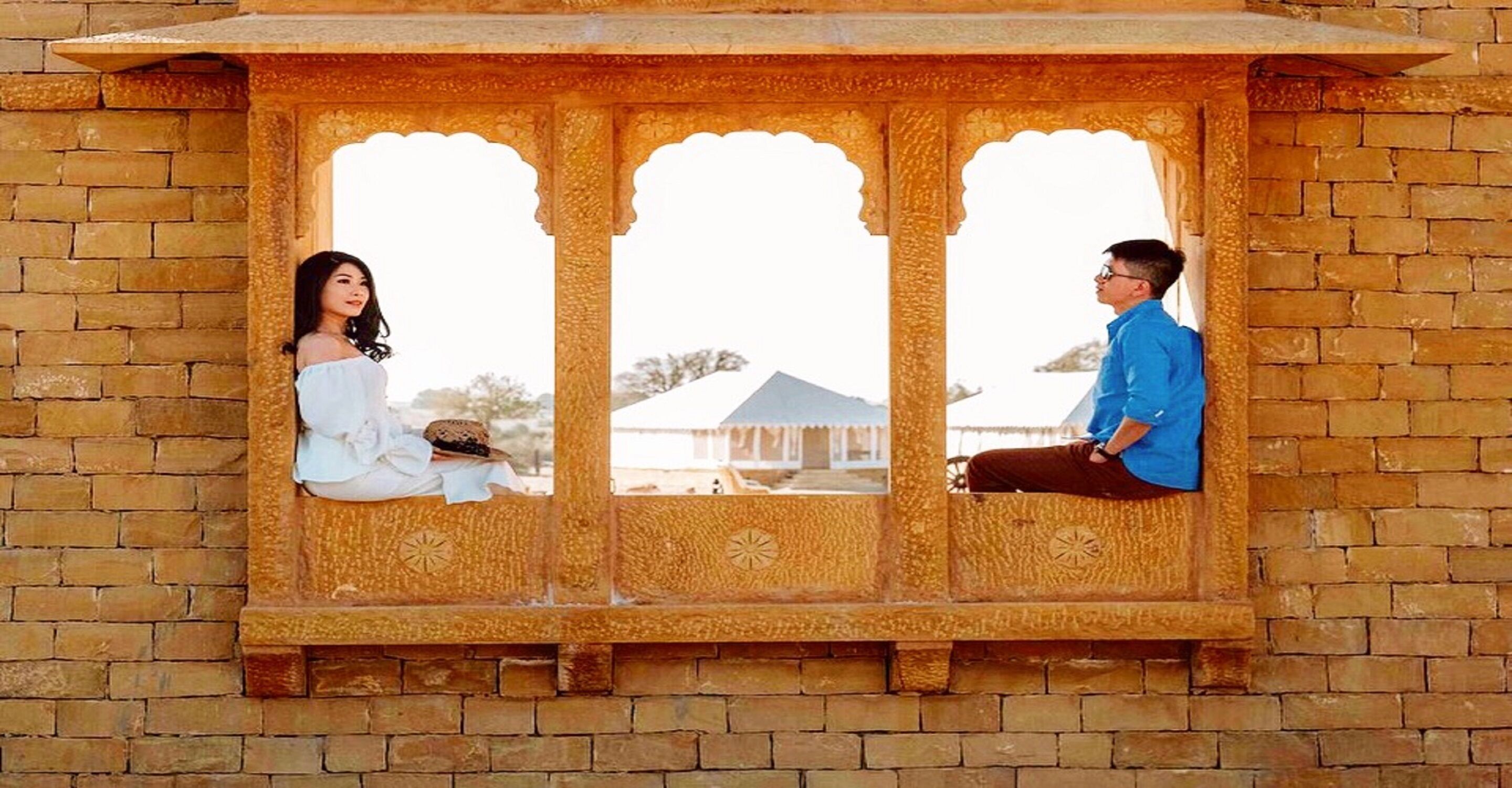 A couple sitting in a windowed alcove with intricate carvings, overlooking a building with a traditional design.