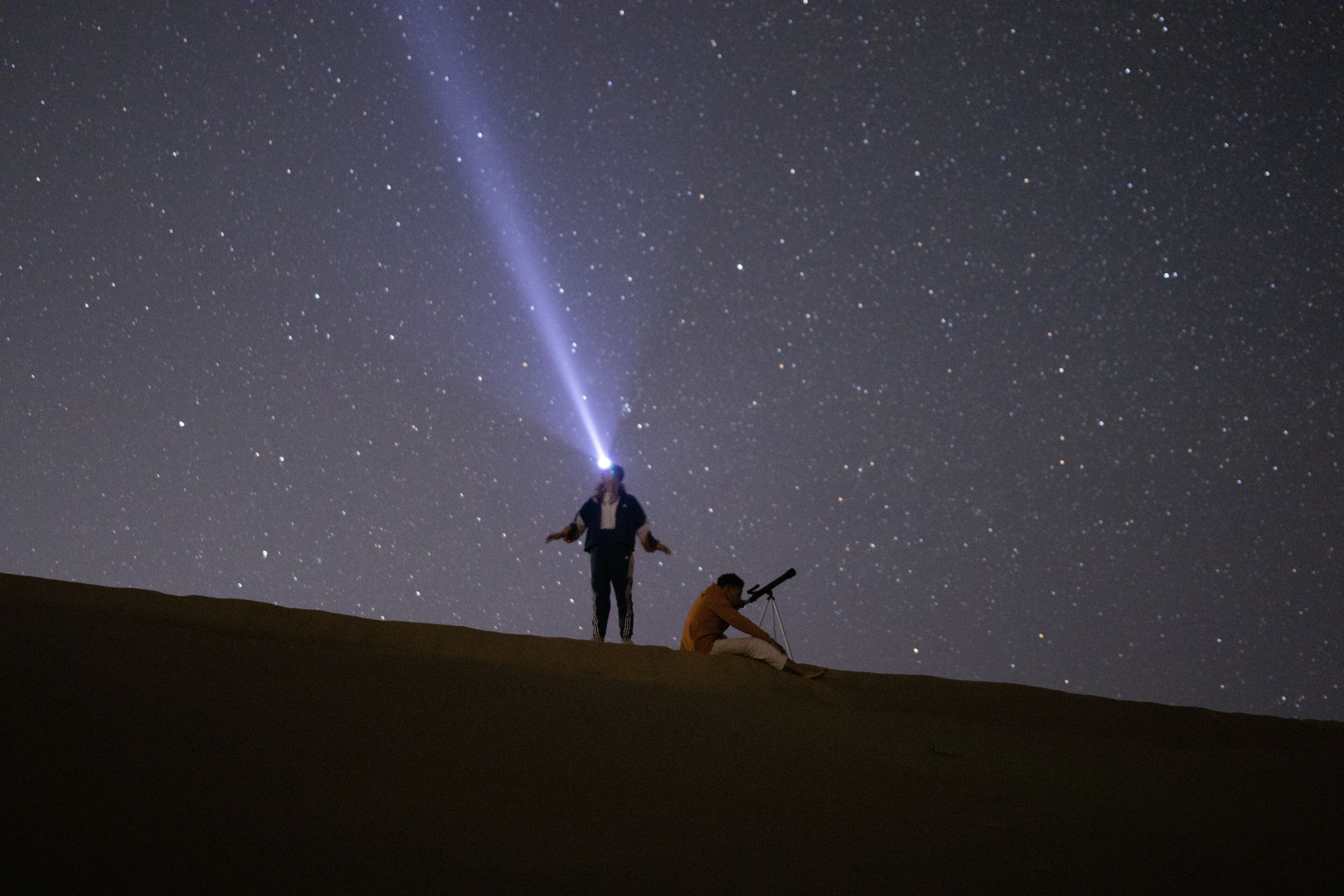 A desert landscape at night with a person standing and another person kneeling with a telescope.