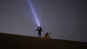 A desert landscape at night with a person standing and another person kneeling with a telescope.