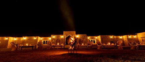 Un patio con mobiliario exterior y una chimenea central, rodeado de muros de piedra e iluminado con luz cálida.