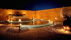 Un espace extérieur entouré de murs en pierre, agrémenté d’une fontaine circulaire, de sièges et de bougies.