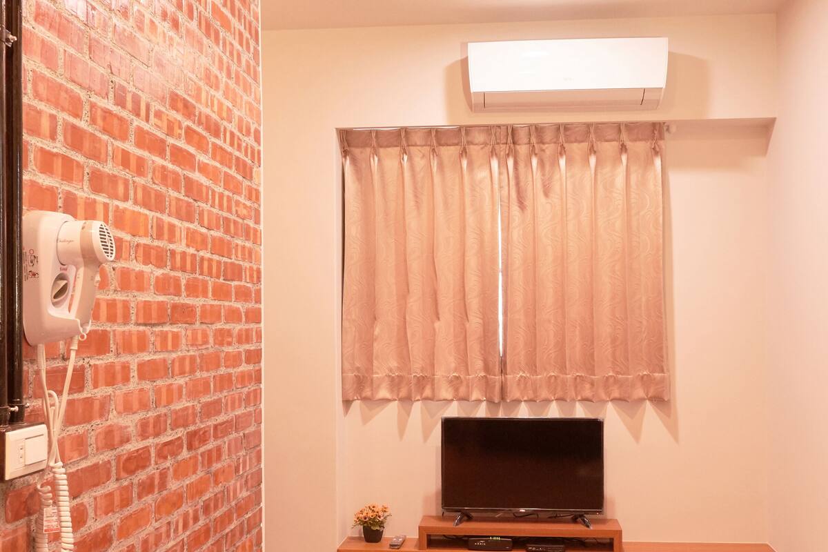 A room with a brick wall, a mounted TV, a ceiling light, and a window with curtains.
