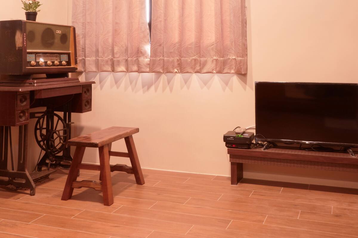 A room with a wooden floor, a wooden table, a radio, a television, and a window with curtains.