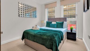 A bedroom with a bed, a bedside table, a wall art piece, and a window with blinds. - MySquare Apartments Shoredtich (London)