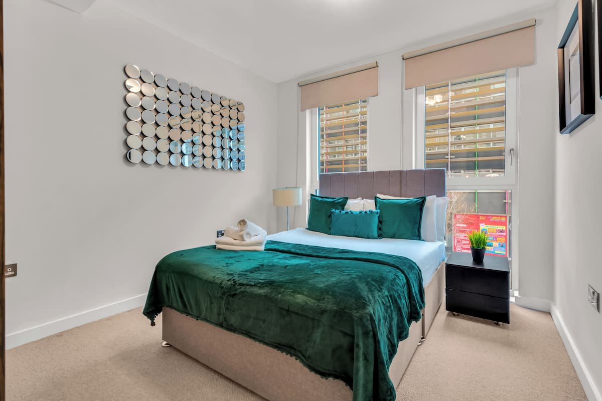 A bedroom with a bed, a bedside table, a wall art piece, and a window with blinds.